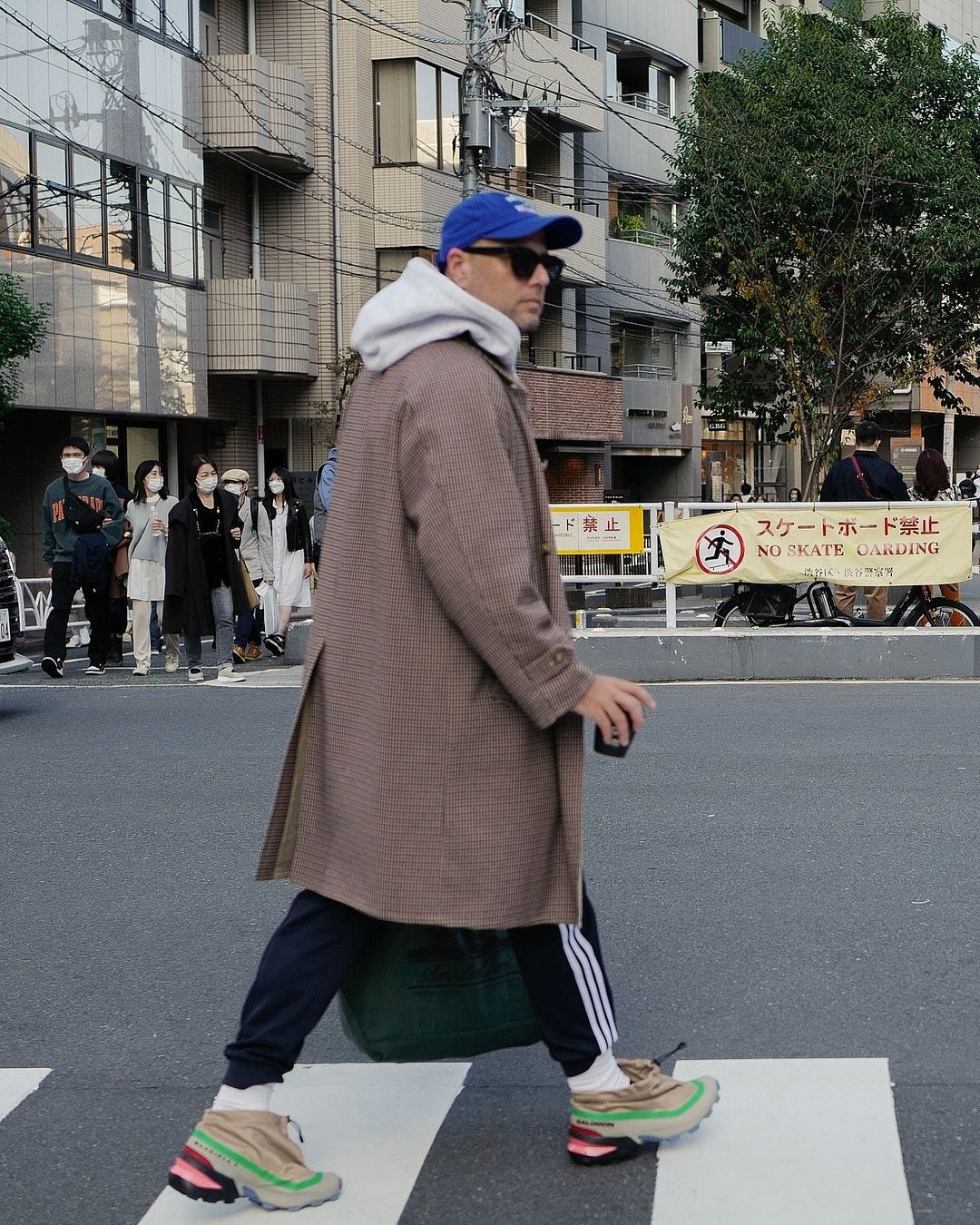 Man crossing the street wearing oversized patterned coat, black sweatpants and gray hoodie holding cup of coffee