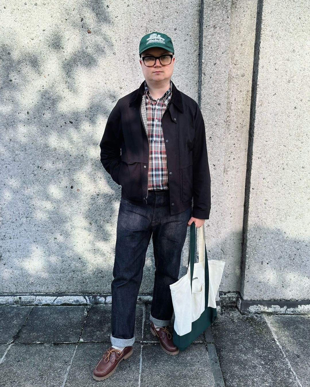 Man standing in front of concrete wall outside wearing navy jacket over plaid madras pop shirt with dark blue jeans, brown moccasins and holding a handbag