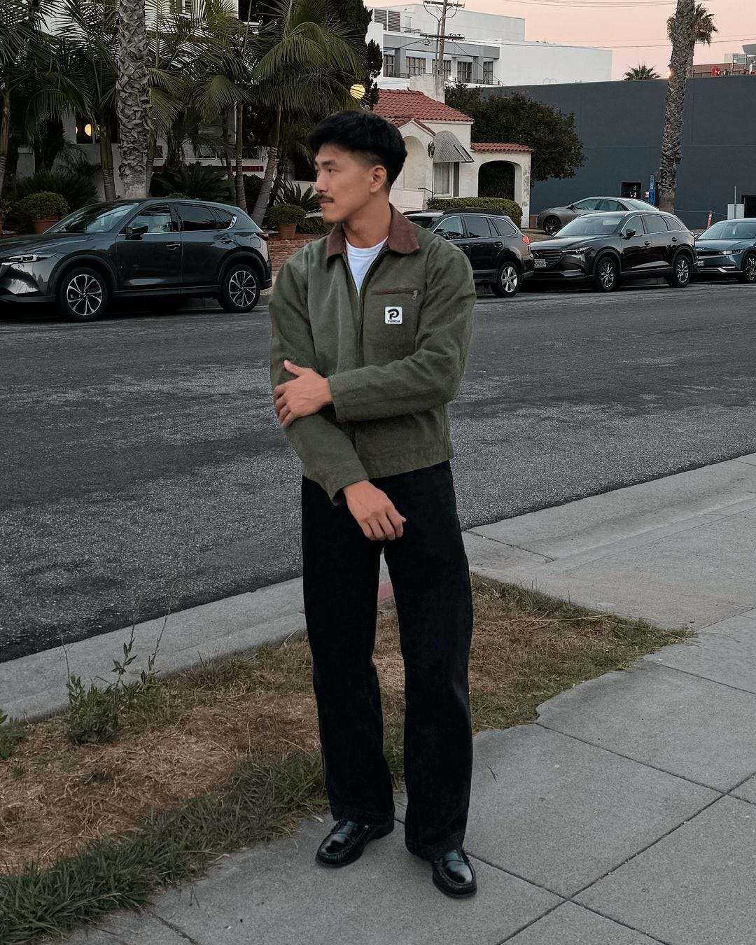 Man standing on suburban street next to driveway wearing olive green brown collared work jacket, white t-shirt, black pants and black loafers