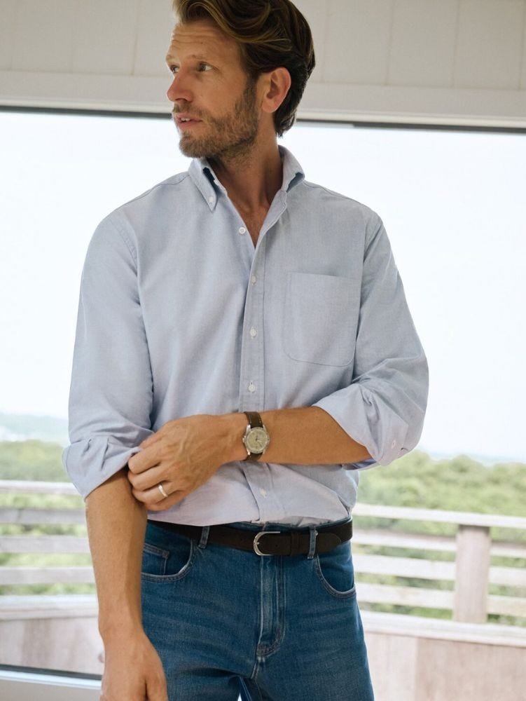 man standing in front of a window wearing a light blue oxford shirt with jeans and a brown belt