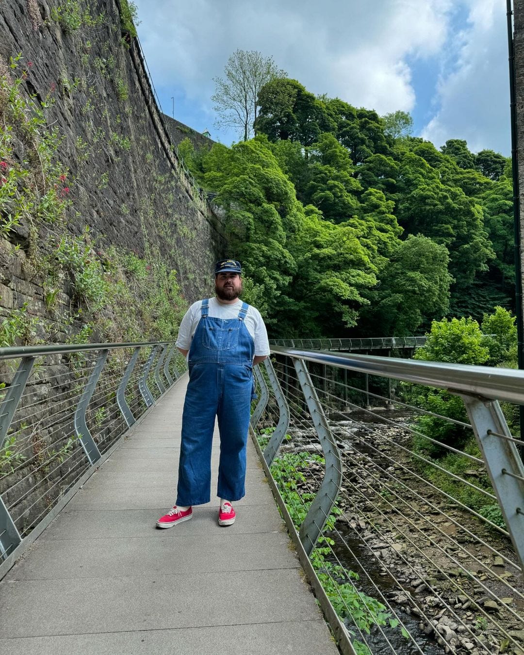 bearded man standing outside on a walkway next to a tall wall and in front of green trees, wearing a baseball hat, a grey t-shirt, blue denim overalls and red canvas sneakers