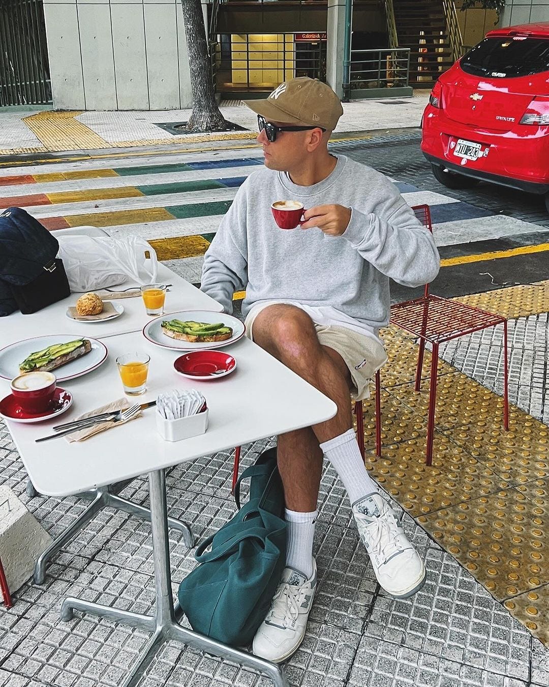 Man sitting in outdoor cafe drinking coffee wearing gray sweatshirt, shorts and white sneakers and high socks
