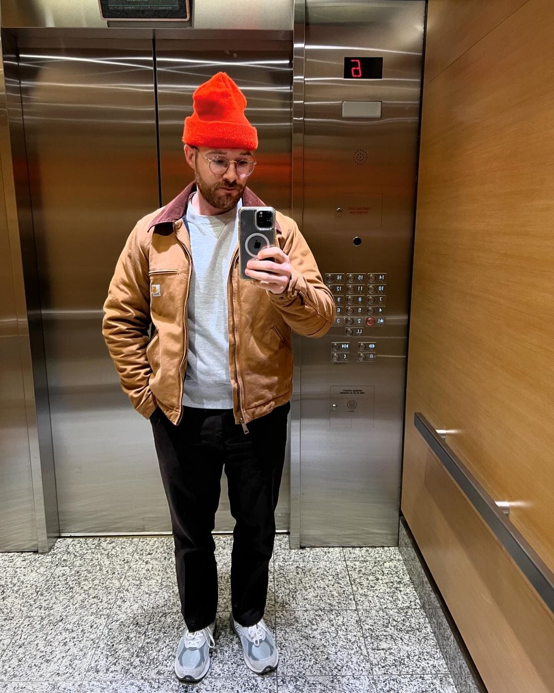 man in elevator taking a selfie wearing dark pants, sneakers, a grey sweatshirt, brown work jacket, and red beanie