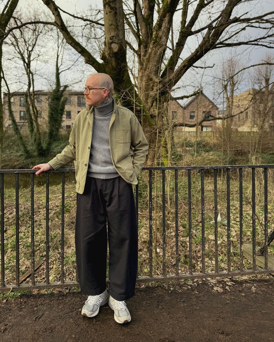 man in park wearing a grey turtleneck, olive military jacket, wide-leg pants, and white sneakers