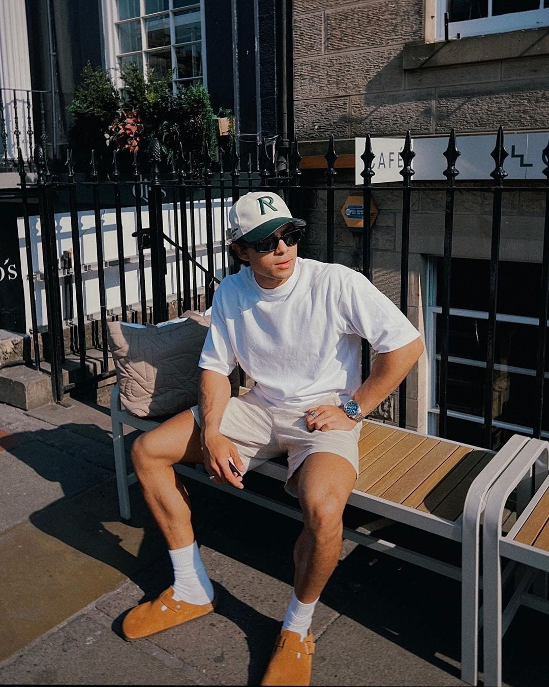 Man sitting on a bench outside wearing a baseball cap, white t-shirt, shorts, high socks and Birkenstocks