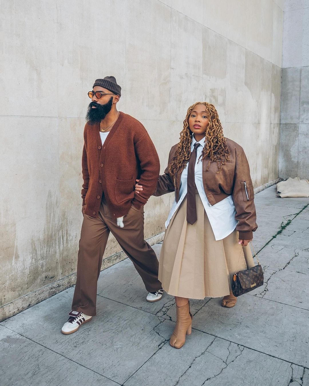 couple walking down the street wearing coordinating brown outfits. the man is in a brown cardigan and brown pants, while the woman is wearing a satin brown jacket and khaki-colored long skirt