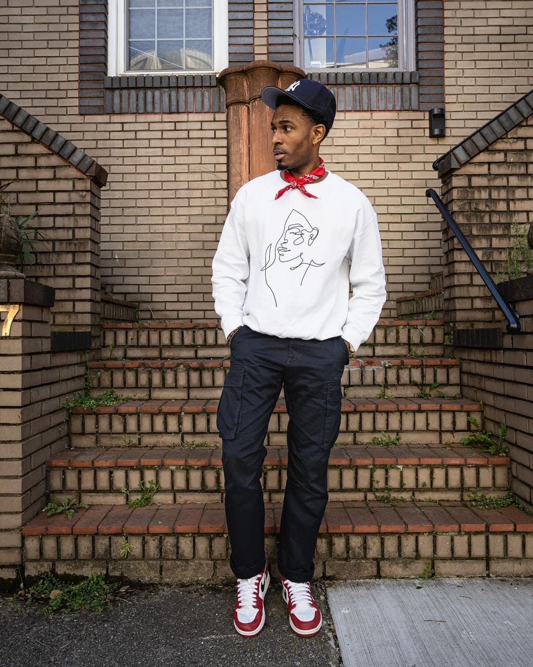 young man in white sweatshirt with red bandana tied around his neck. he's also wearing a dark baseball cap, dark jeans, and white sneakers with red accents