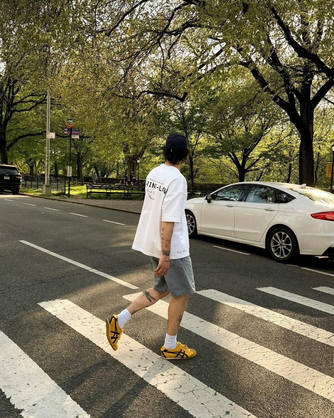 Photo of a man crossing the road with his back to the camera, wearing a white T-shirt, denim shorts, high white socks and yellow sneakers