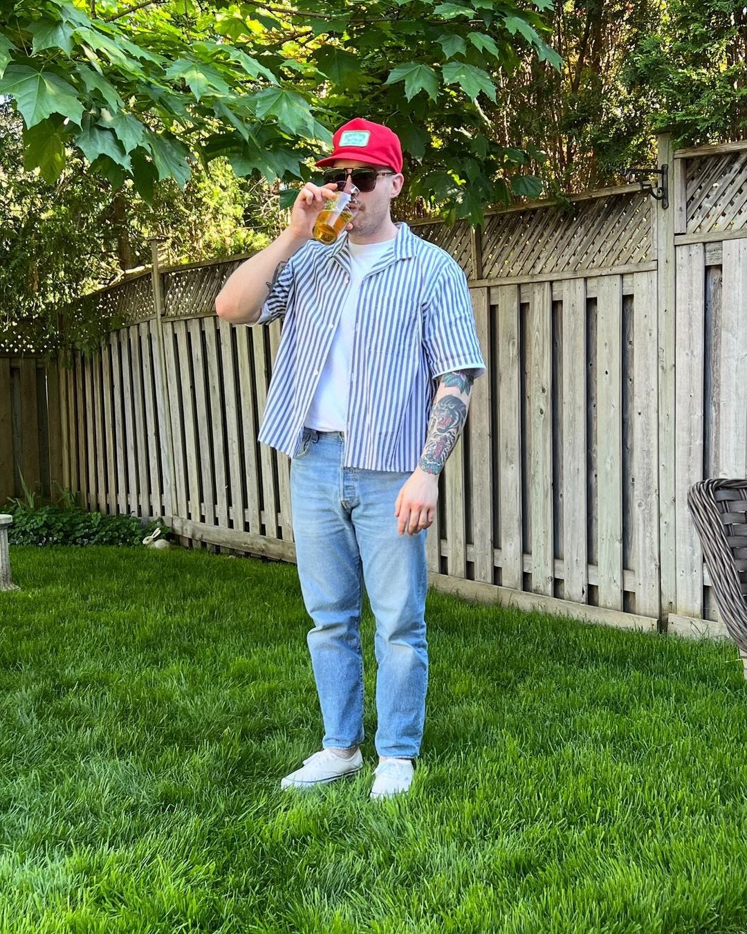 man standing in a backyard in front of a frence, wearing a red cap, blue striped short sleeve shirt, jeans, and white sneakers, drinking a beer
