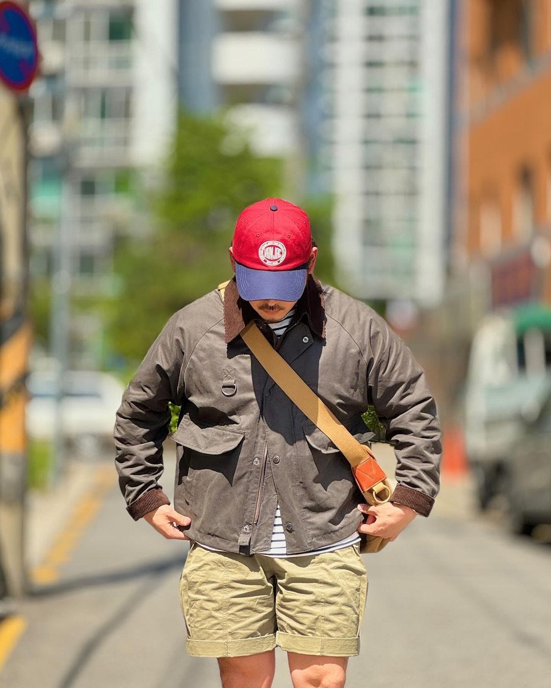 man in red baseball cap with blue bill, olive-colored jacket, brown bag across his body, chino shorts, tall white socks, and red sneakers