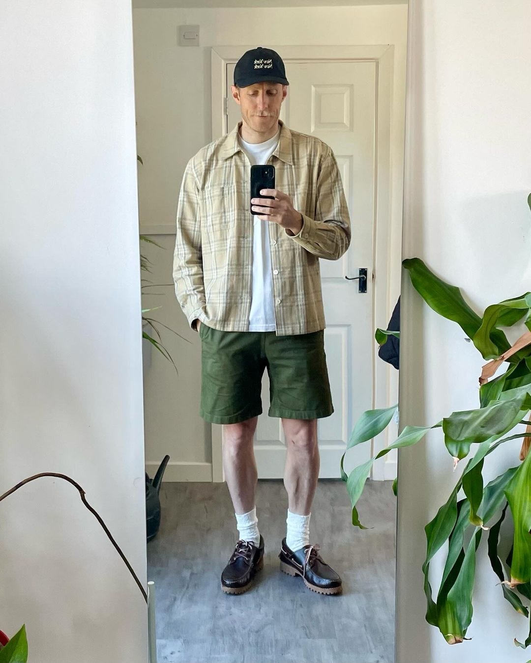 man taking a picture of himself in the mirror. he is wearing a long-sleeve tan shirt over a white t-shirt, green shorts, and boat shoes with tall white socks