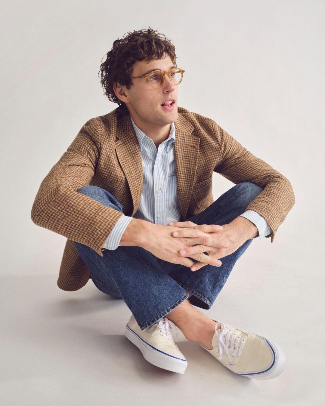 man wearing a blazer and jeans with white canvas sneakers sitting on the floor with his legs crossed