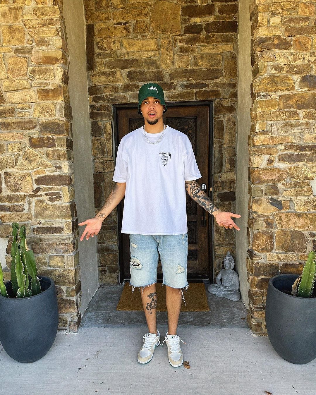 The man standing at the door is wearing a green baseball cap, a white T-shirt, light-colored ripped denim shorts and Nike sneakers