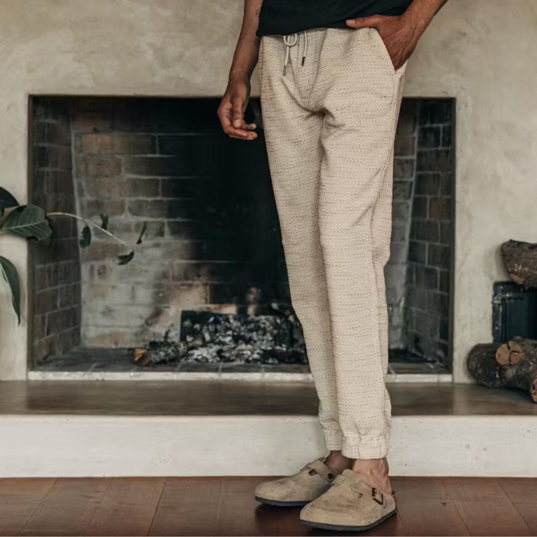 lower half of a man standing in front of a fireplace wearing cream-colored sweatpants with suede Birkenstock clogs on his feet
