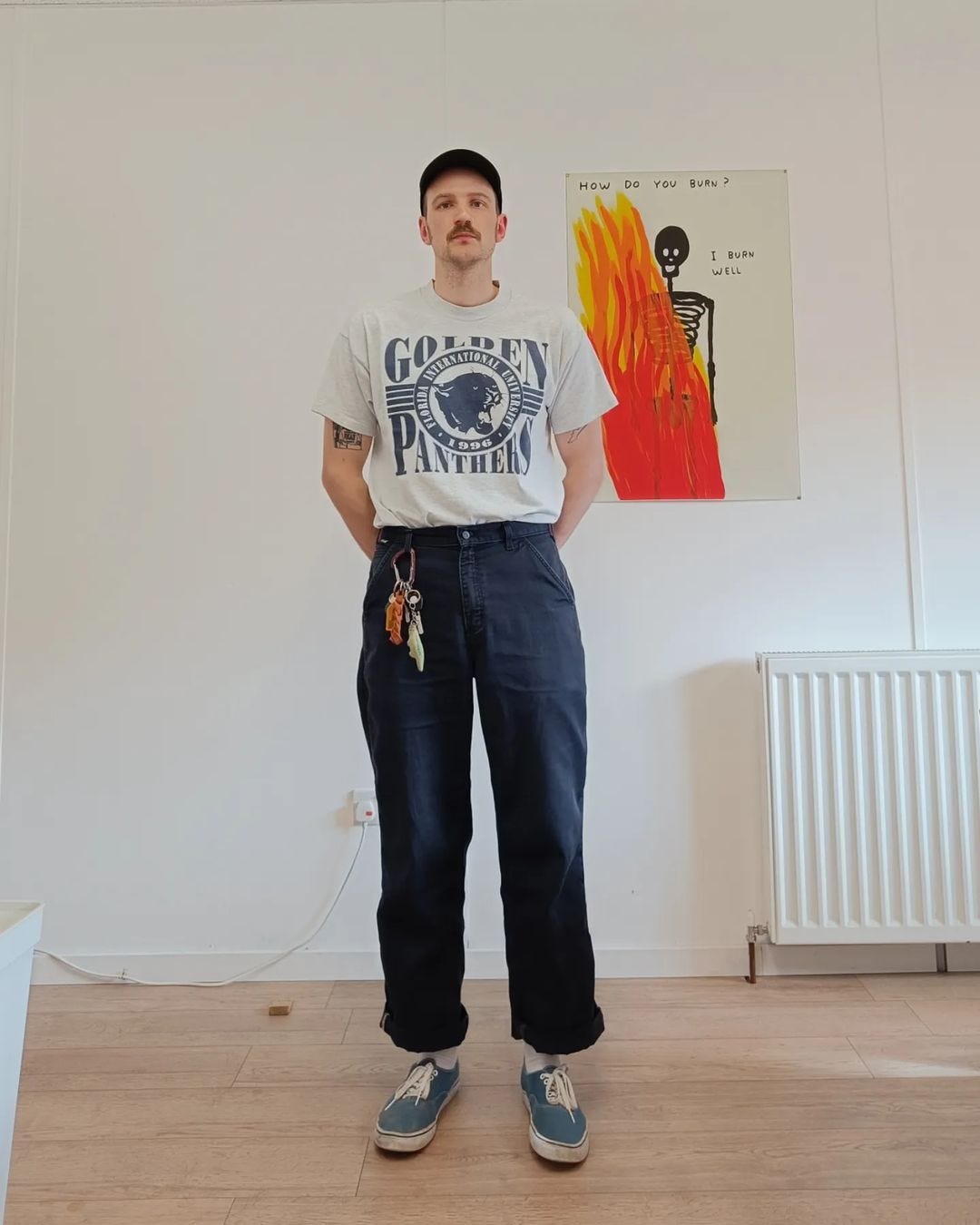 man standing in front of a white wall with a bright-colored painting on it, wearing a graphic t-shirt with dark pants and blue sneakers