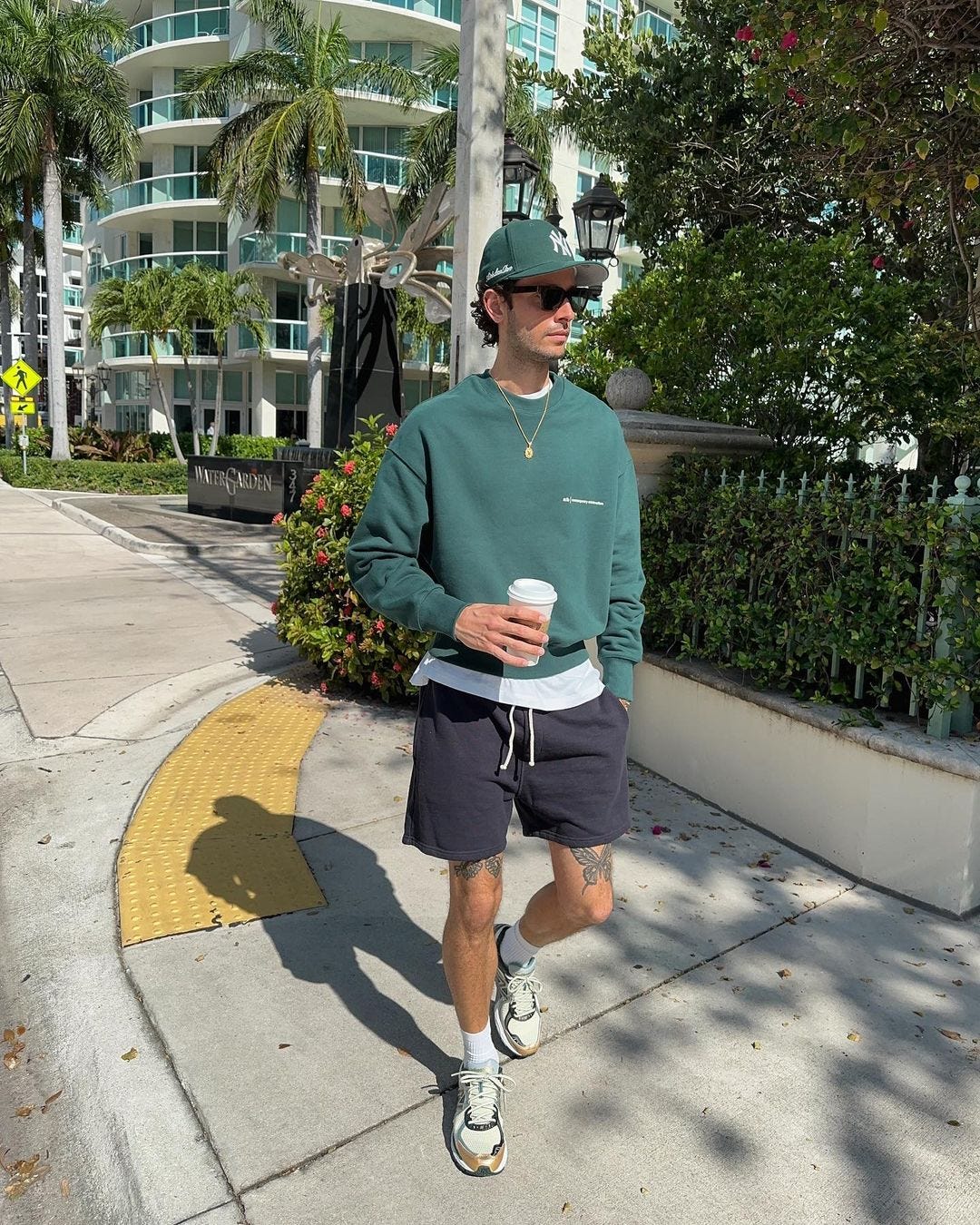 man in a green baseball cap, green sweatshirt, navy shorts, and white sneakers, holding a coffee cup