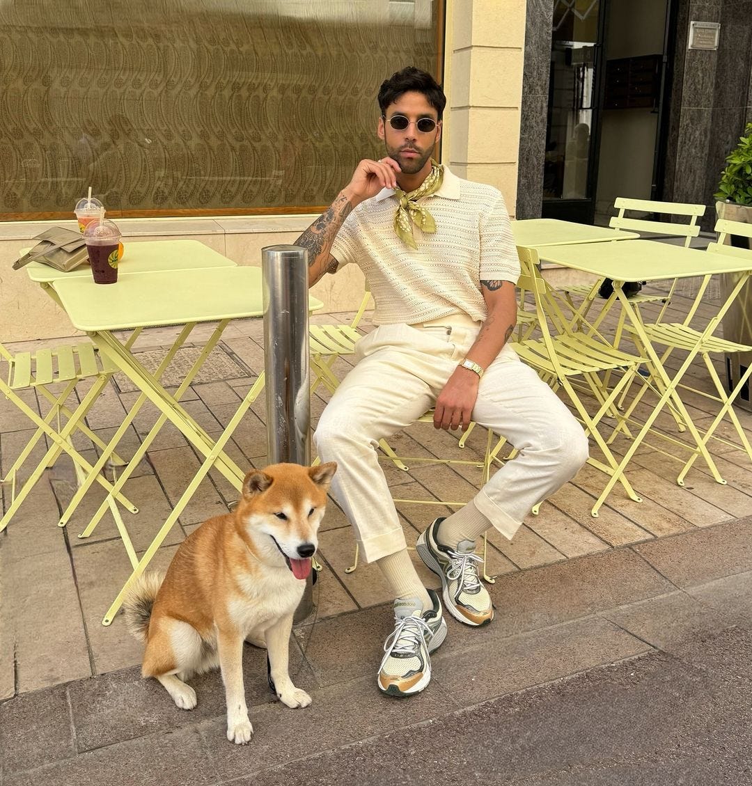 man sitting at a cafe table outside, wearing a white crochet top, white pants, a light green bandana around his neck and sunglasses. a dog is at his feet | light colored outfits for men