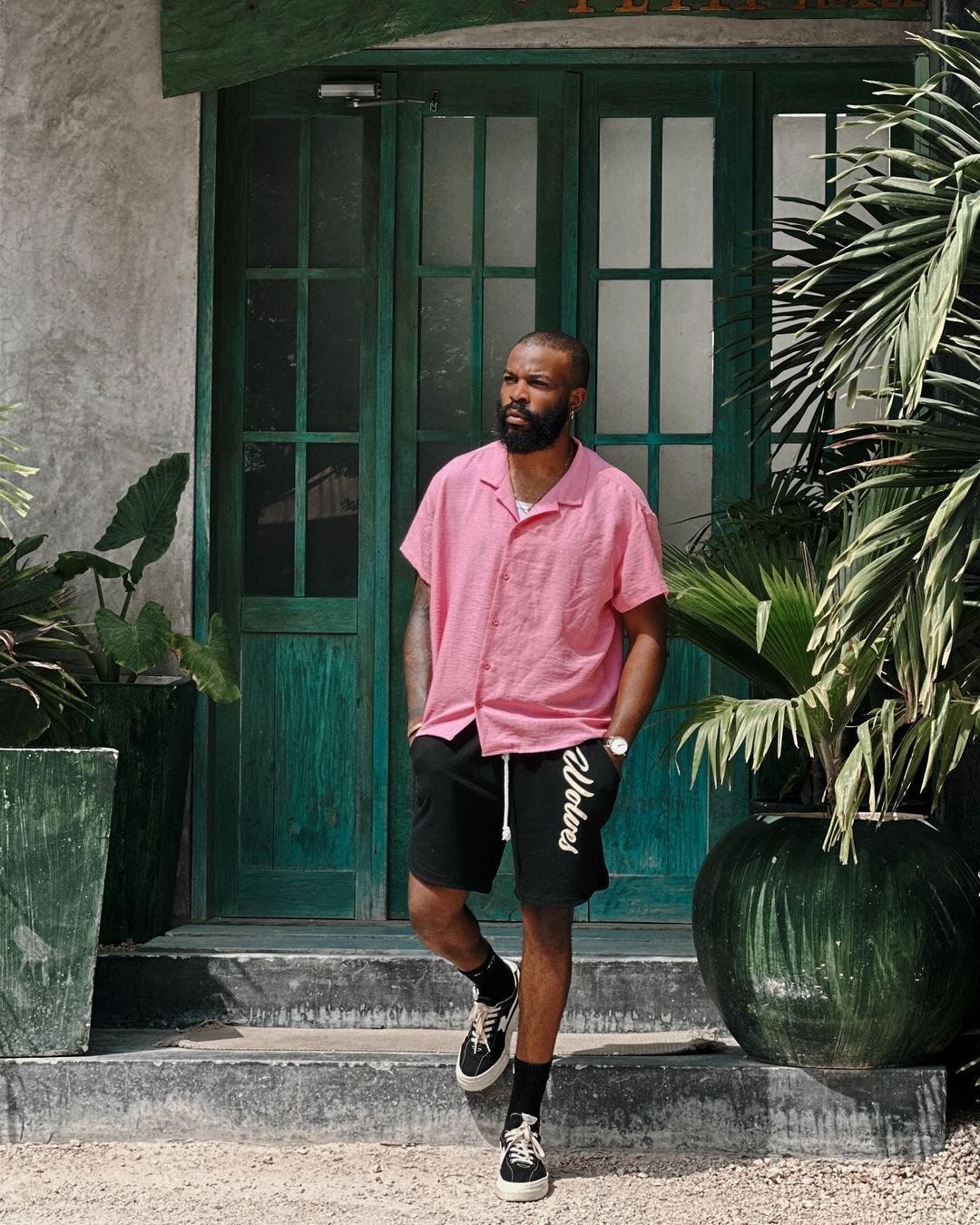 man standing outside on steps in front of a green door in pink short sleeve button down shirt, black shorts with a white drawstring, and black sneakers