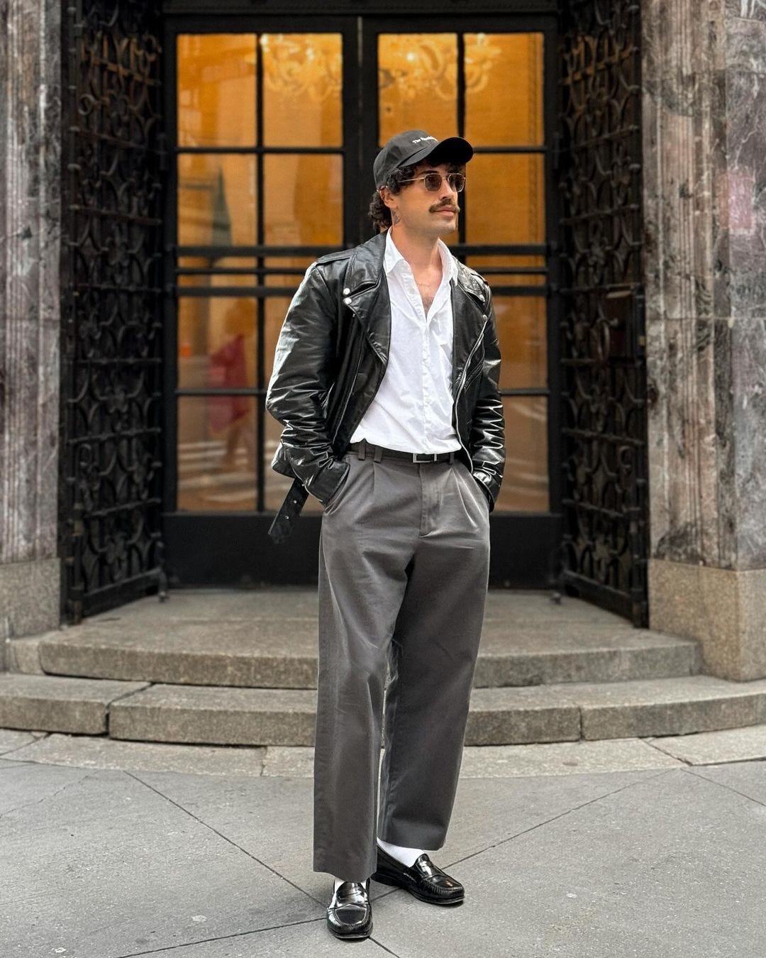 A man stands in front of a building wearing a black leather jacket, white button-down shirt, gray slacks, black loafers and white socks