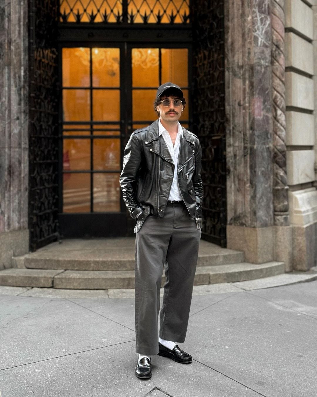 man standing on a street corner wearing a leather jacket, grey dress pants, and black loafers with white socks