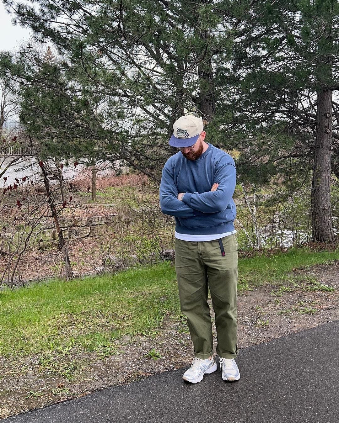 man standing outside with his arms folded across his chest and looking down. he's wearing a baseball cap, blue sweatshirt, olive-colored chinos and white sneakers