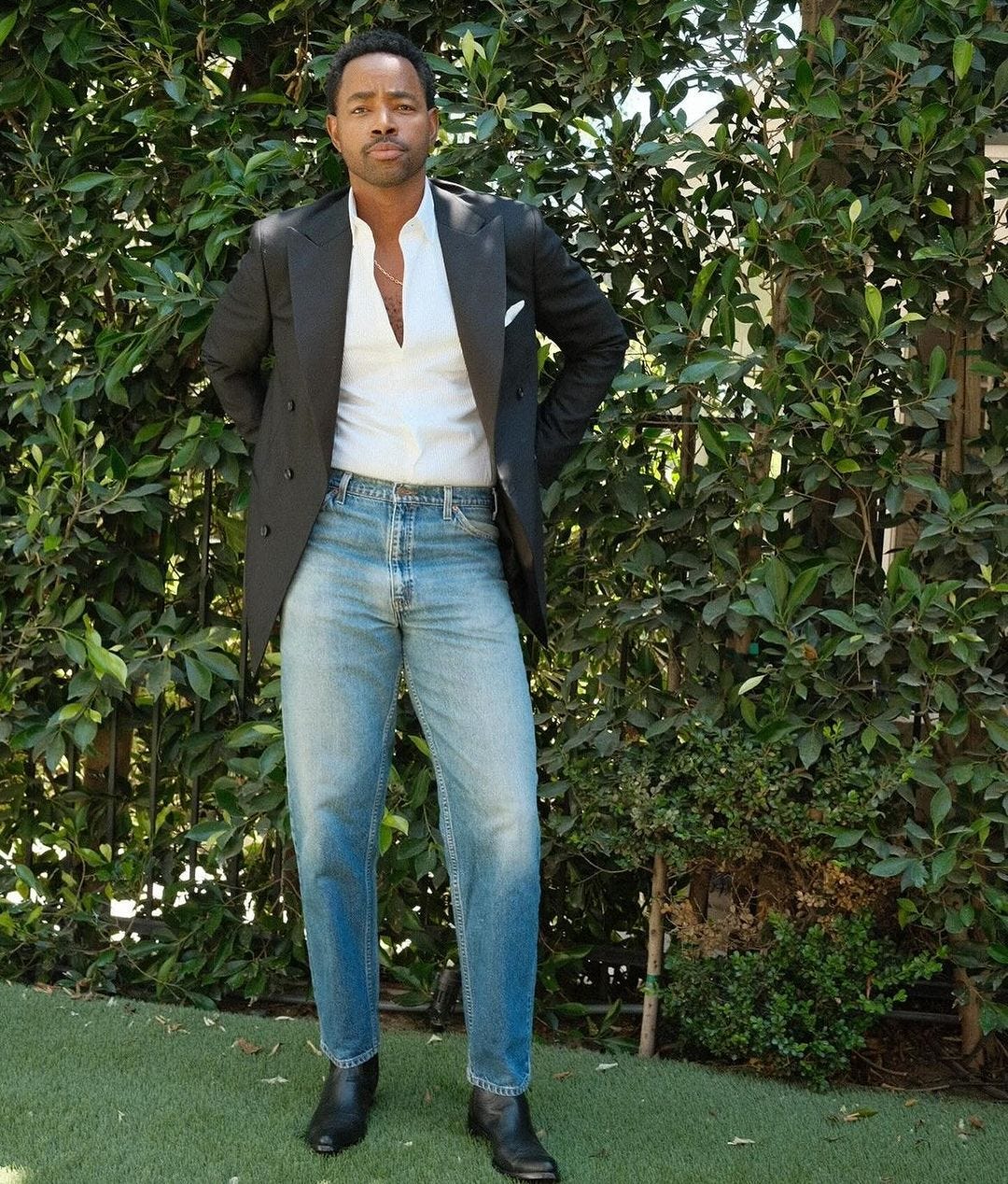 Jay Ellis wearing a black tuxedo jacket and light wash jeans, standing in front of a wall of plants