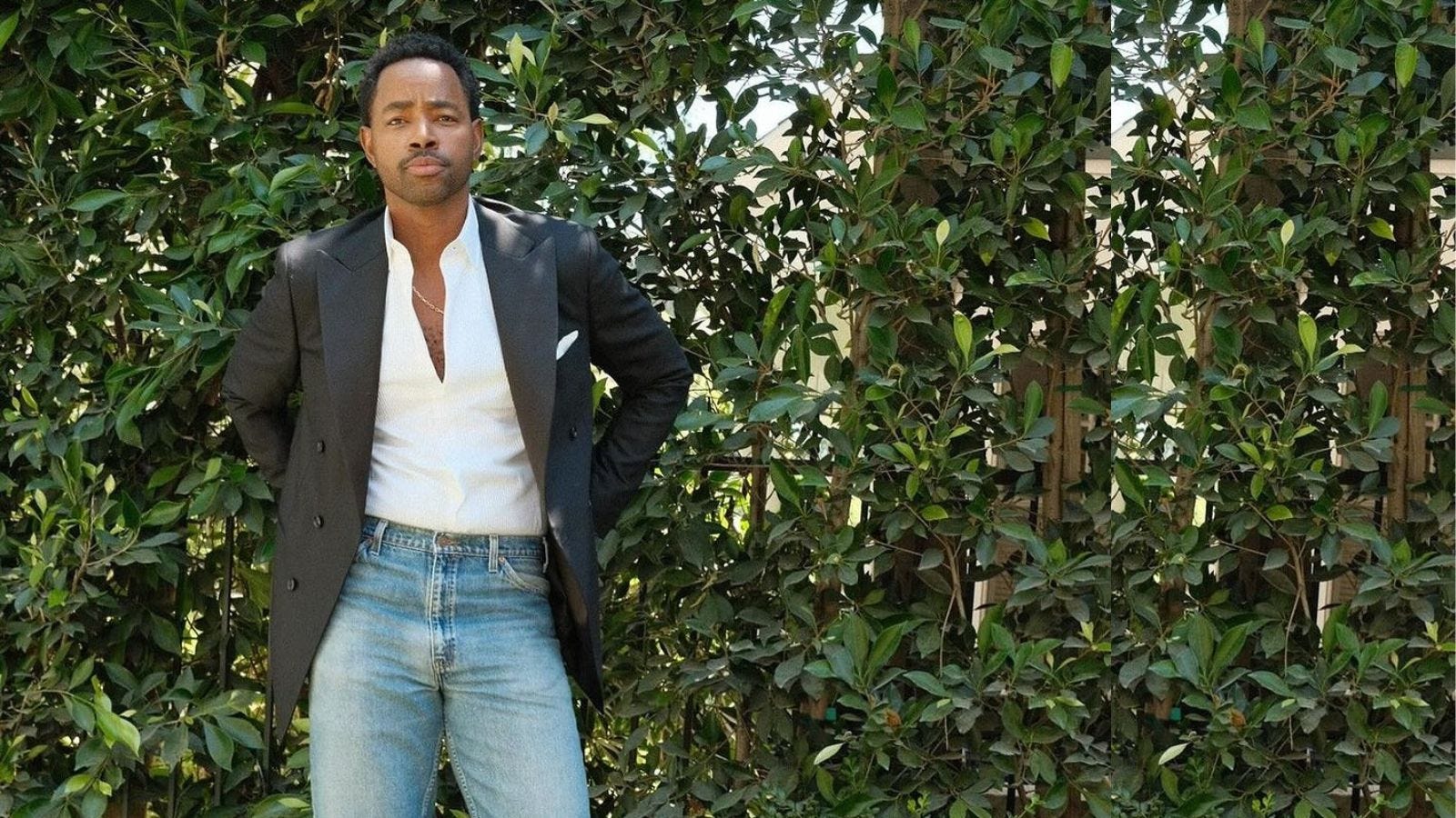 actor Jay Ellis wearing a black tuxedo jacket, white shirt, and light wash jeans, standing against a wall of greenery