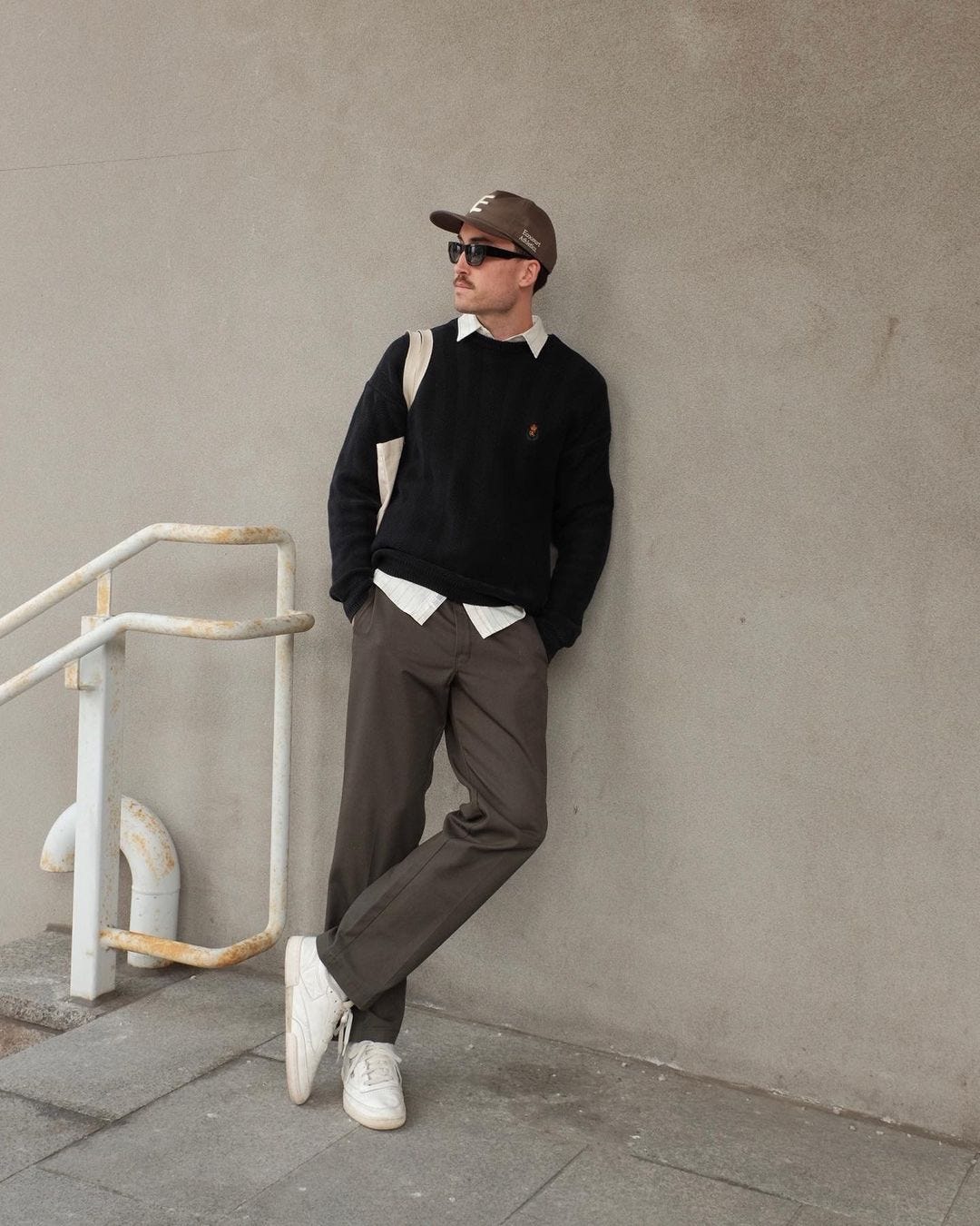 man leaning against a wall wearing sunglasses, a black sweater, dark pants, and white sneakers