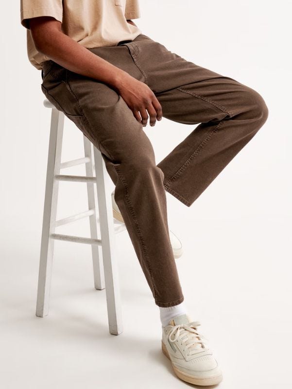 man from the waist down, sitting on a stool, wearing brown workwear pants with white sneakers