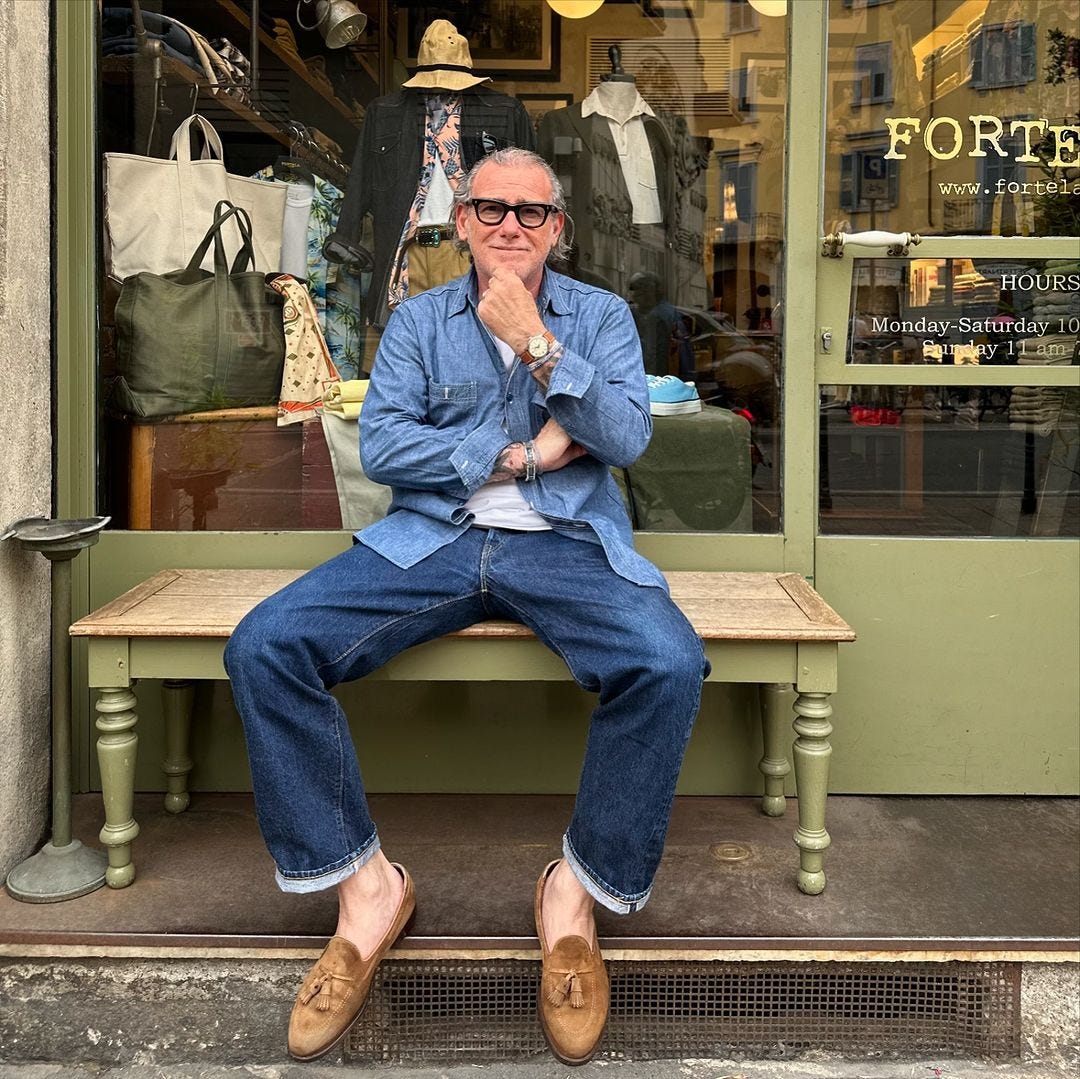 older man sitting outside a retail store wearing a chambray shirt, jeans, and brown loafers