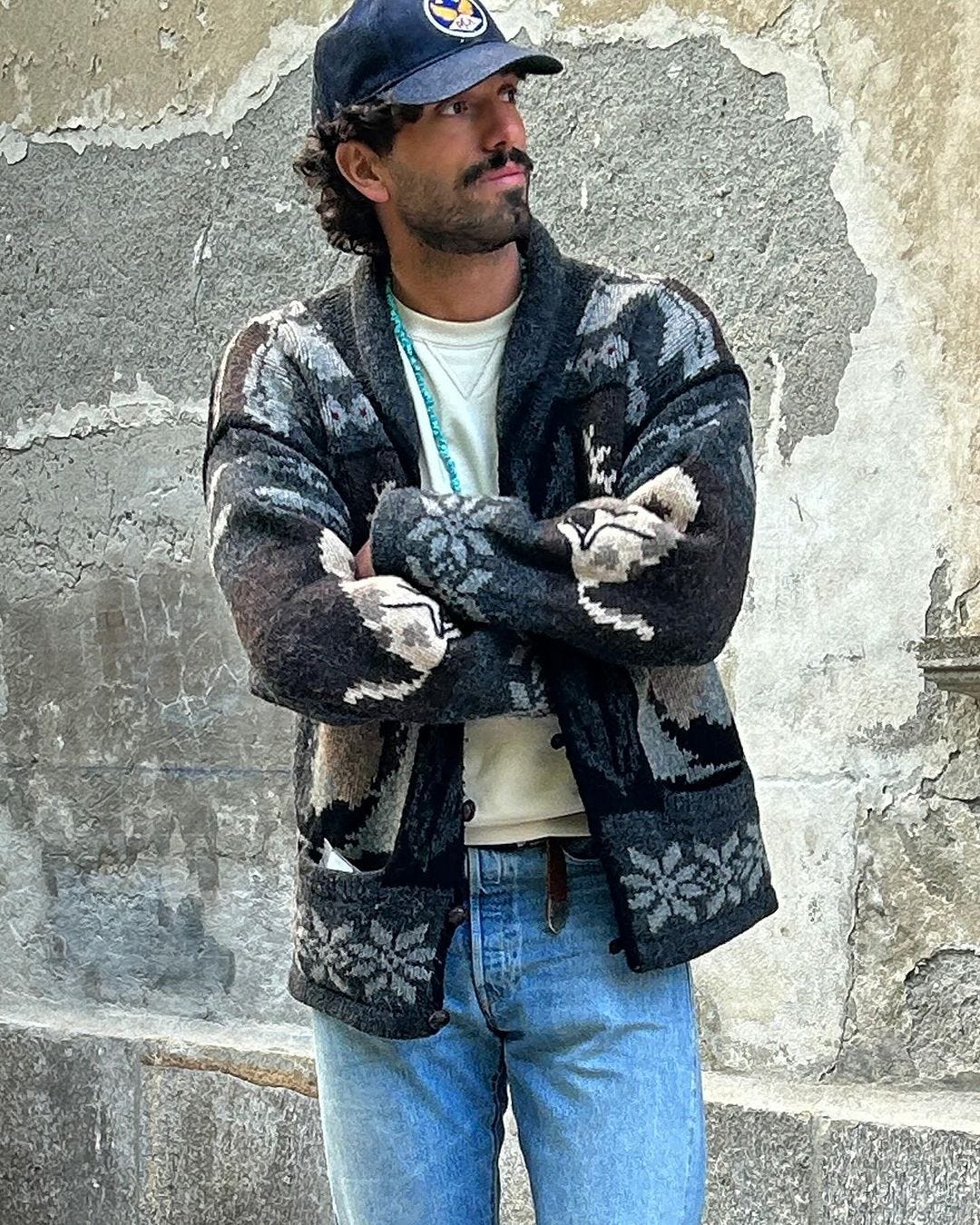 close-up of a man wearing a southwestern-style cardigan with a turquoise necklae, blue baseball cap, cream-colored crewneck shirt, and light jeans