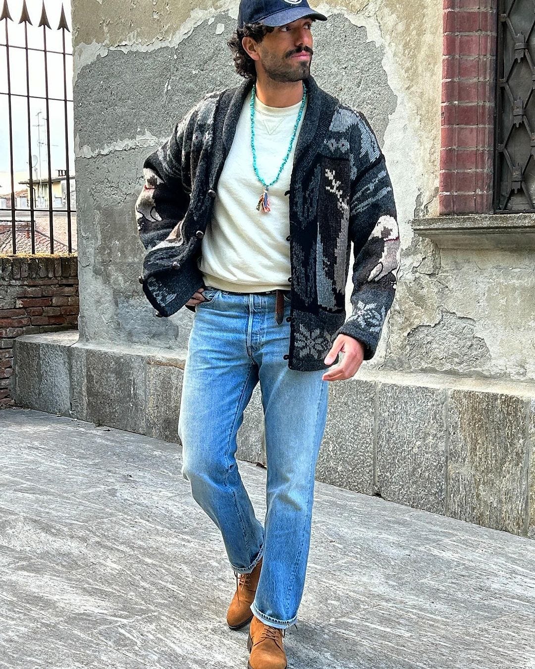 man wearing a southwestern-style cardigan with a turquoise necklae, blue baseball cap, cream-colored crewneck shirt, light jeans and brwon boots