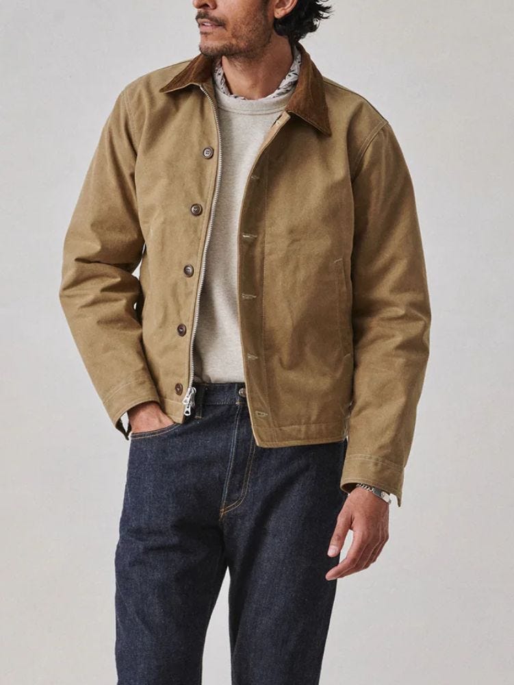 man from the neck down wearing a tan canvas work jacket over a cream sweater and blue jeans