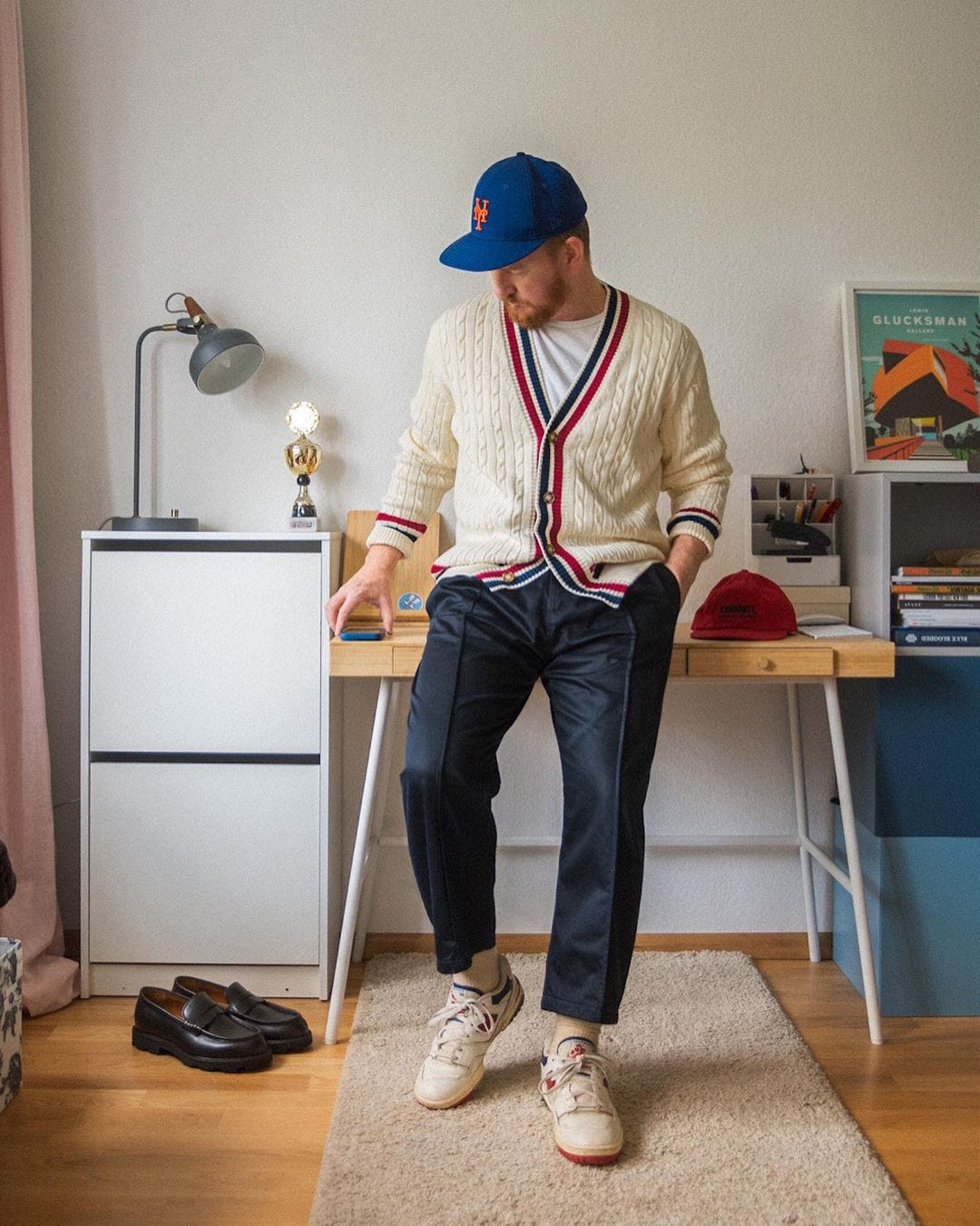 man leaning against a desk, looking down, wearing a blue baseball cap, off-white cardigan with blue and white stripes, dark pants and sneakers