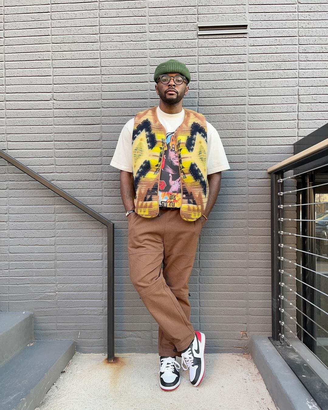 man standing outside wearing a green beanie, white t-shirt with a multi-color vest, brown workwear pants, and nike sneakers