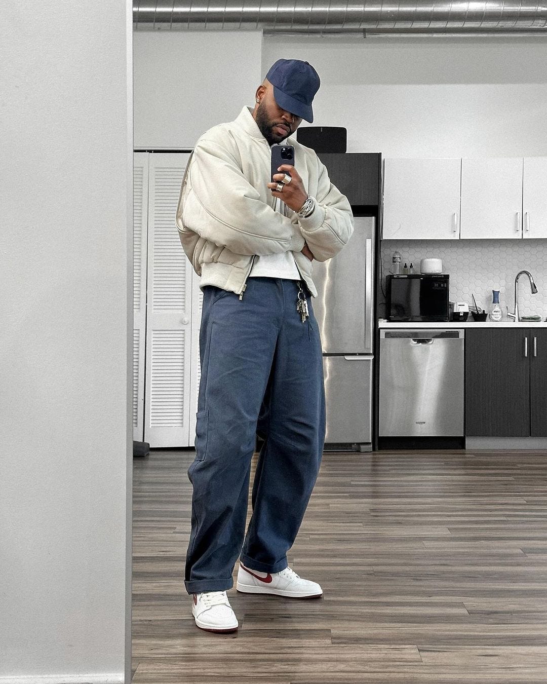 a man taking a selfie in a kitchen, wearing a navy baseball cap, off-white jacket, blue workwear pants, and white Nike sneakers