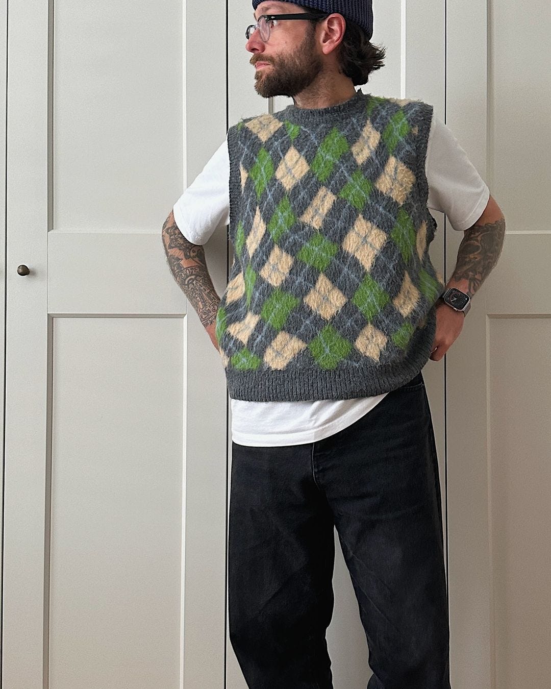 Close-up of a man wearing glasses standing in front of a closet door wearing a patterned green and yellow sweater vest over a white t-shirt and dark pants