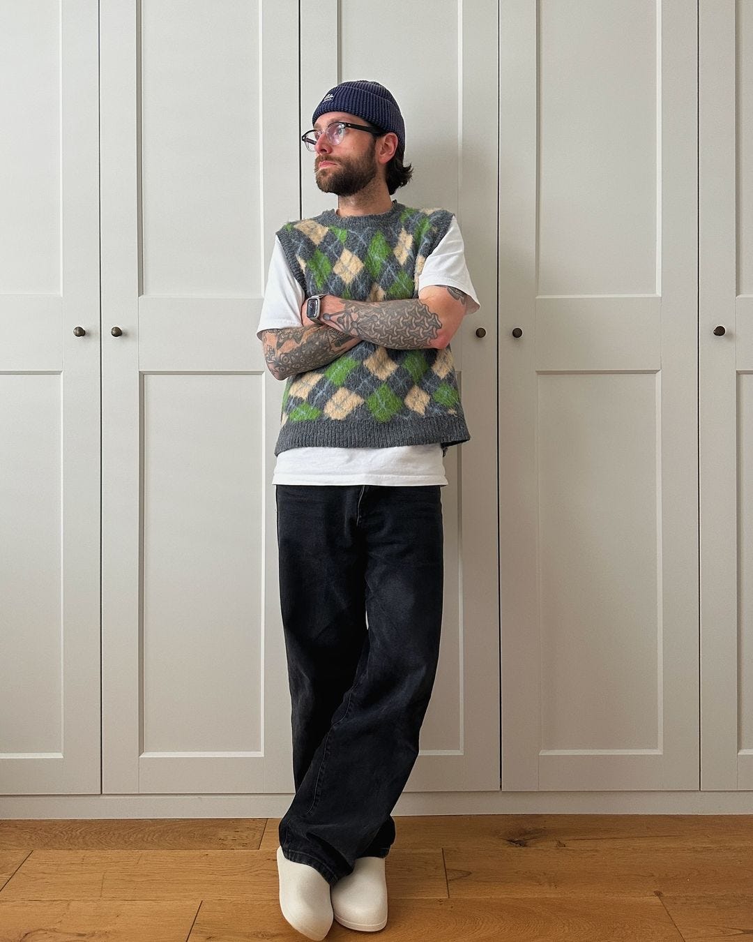 Man wearing glasses standing in front of closet door wearing a patterned green and yellow sweater vest over a white t-shirt and dark pants