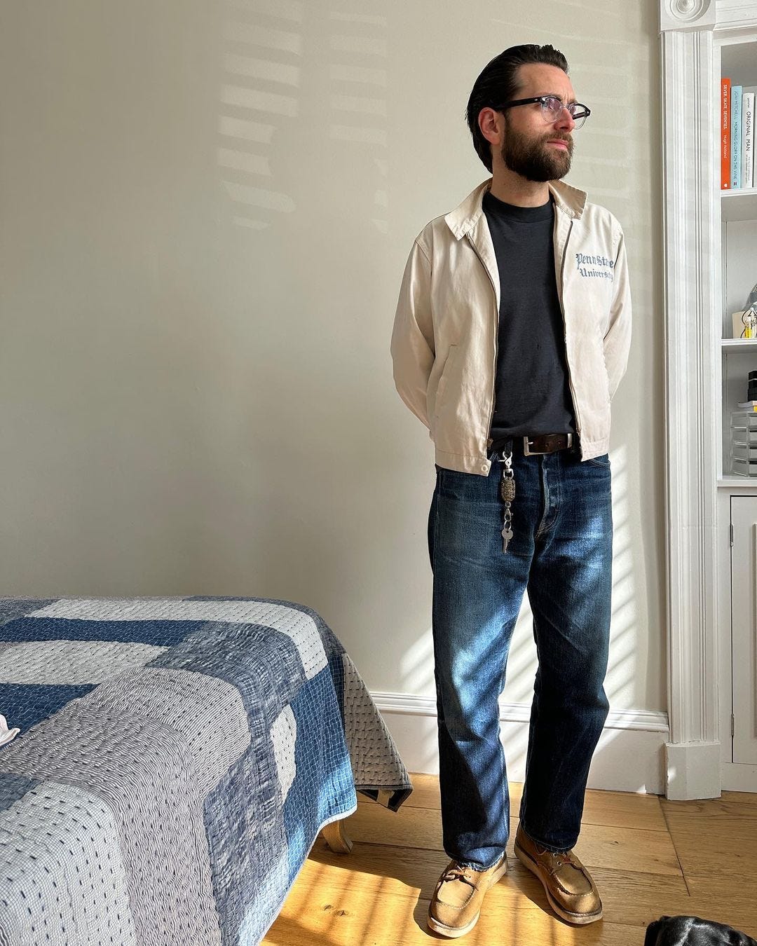 man in a bedroom wearing a cream-colored jacket with Penn State University on the chest over a black t-shirt and dark jeans