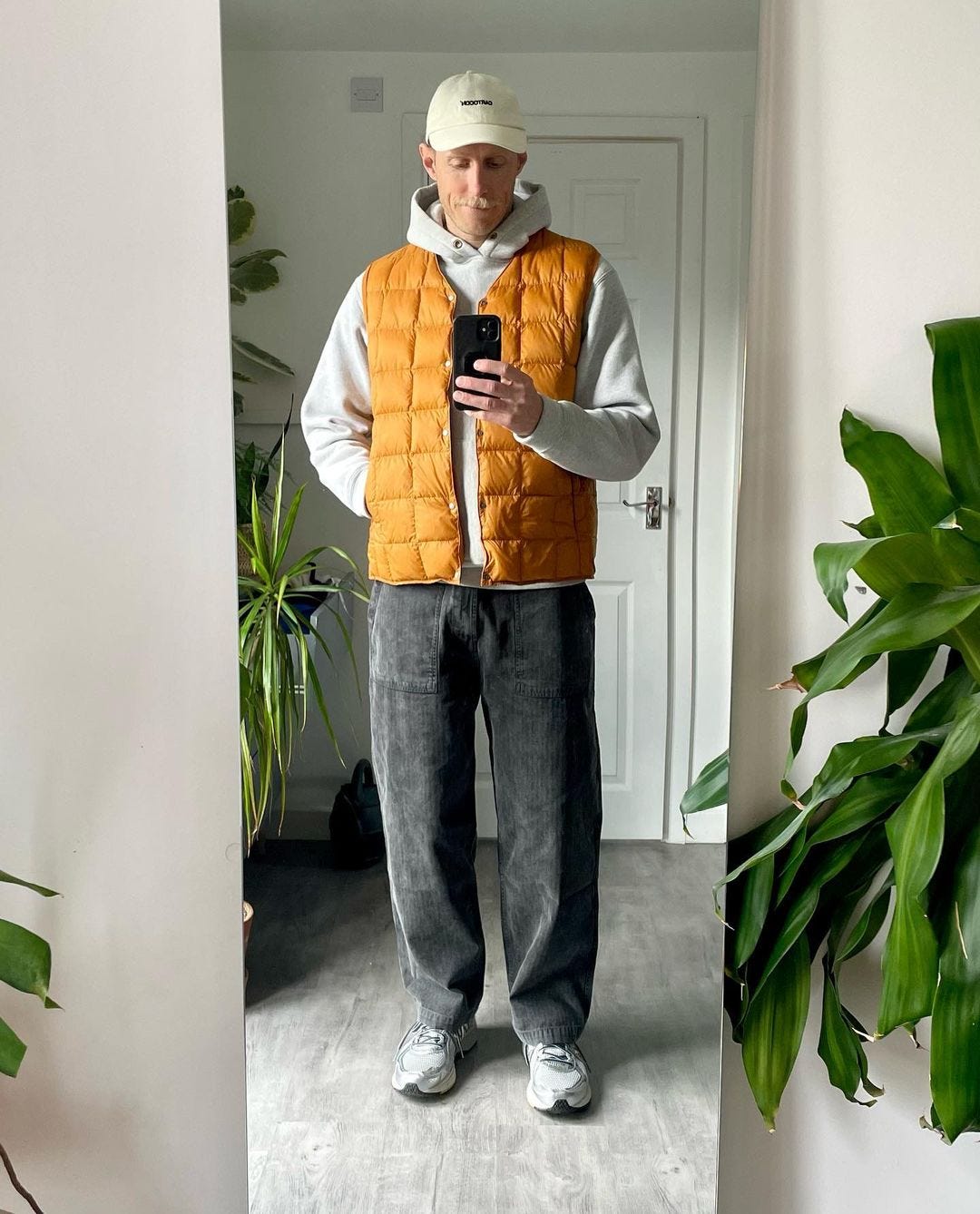 man taking a selfie in a mirror wearing a grey hoodie with an orange quilted vest, grey carpenter pants, and grey sneakers