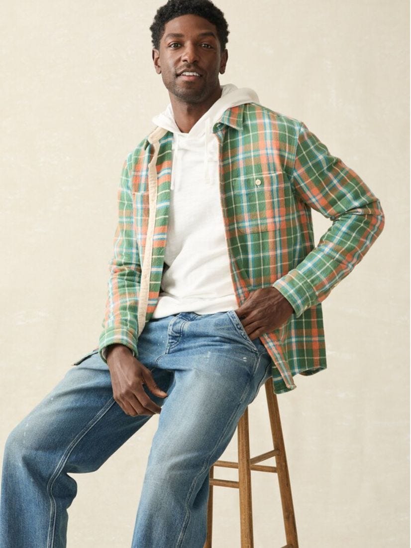 man sitting on a stool wearing a green flannel plaid shirt, cream-colored hoodie, and blue jeans