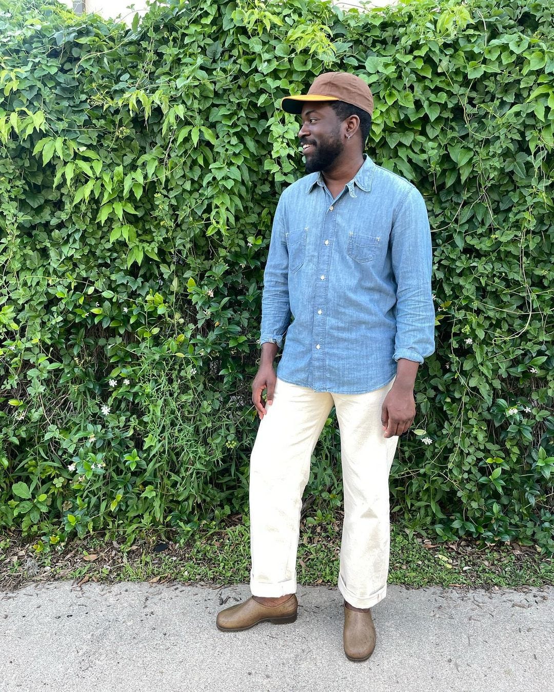 man standing in front of a wall of greenery wearing a chambray shirt and off-white jeans with gardening clogs