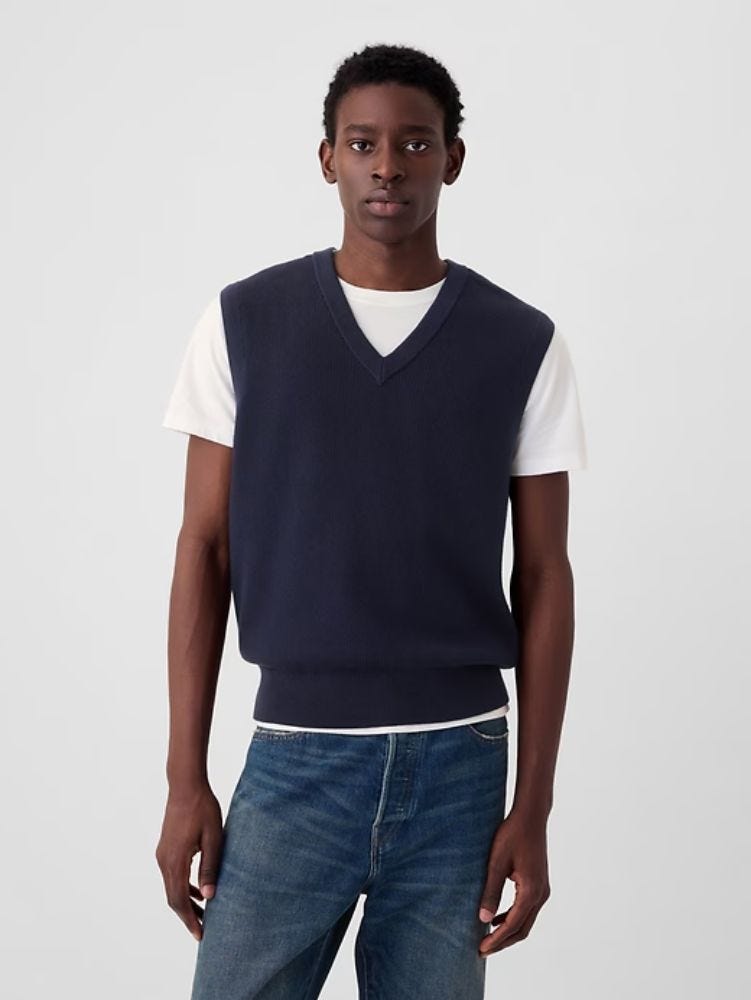 Man wearing navy v-neck sweater vest, white t-shirt and jeans from waist up