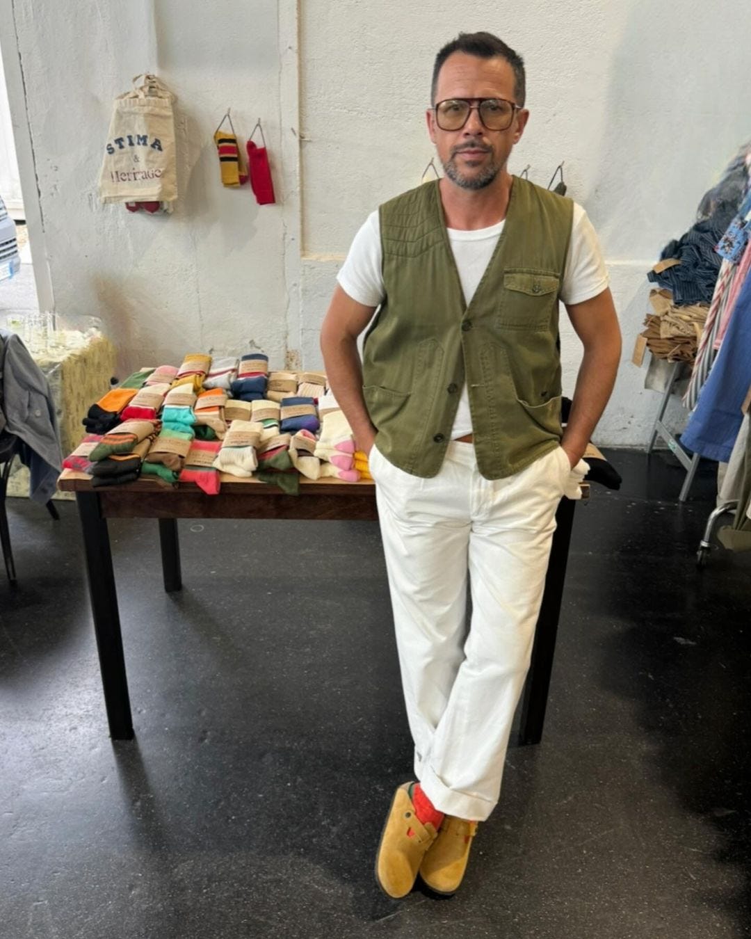 man leaning against a table full of socks, wearing an olive green vest with tan pants and suede birkenstock boston clogs with red socks