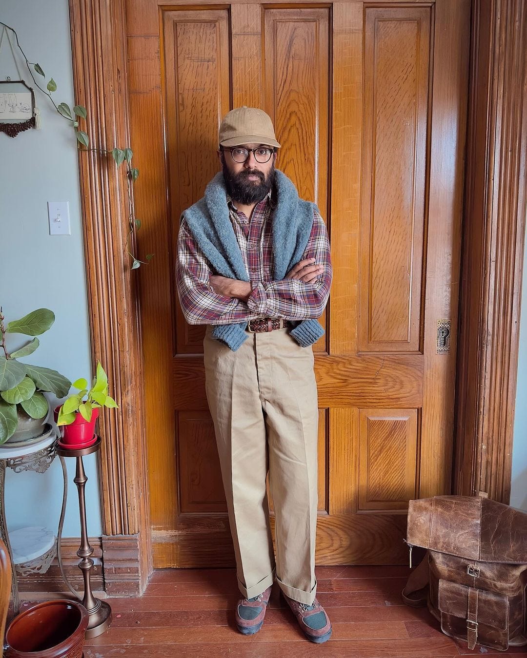 Man standing in front of wooden door, wearing plaid shirt and gray sweater draped over his shoulders