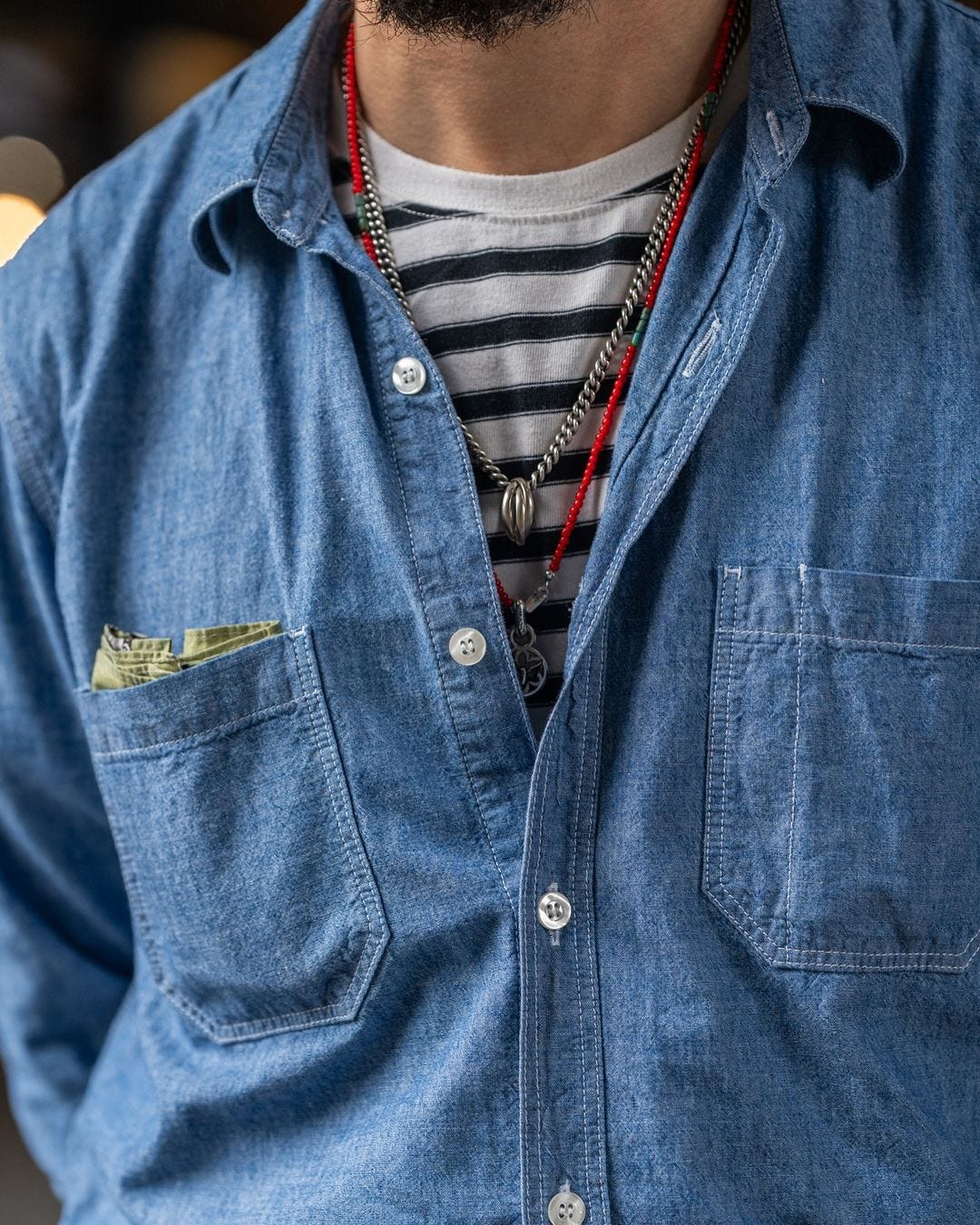 close-up of a man wearing a chambray shirt over a blue and white button-down shirt with two necklaces, and a pocket square peeking out