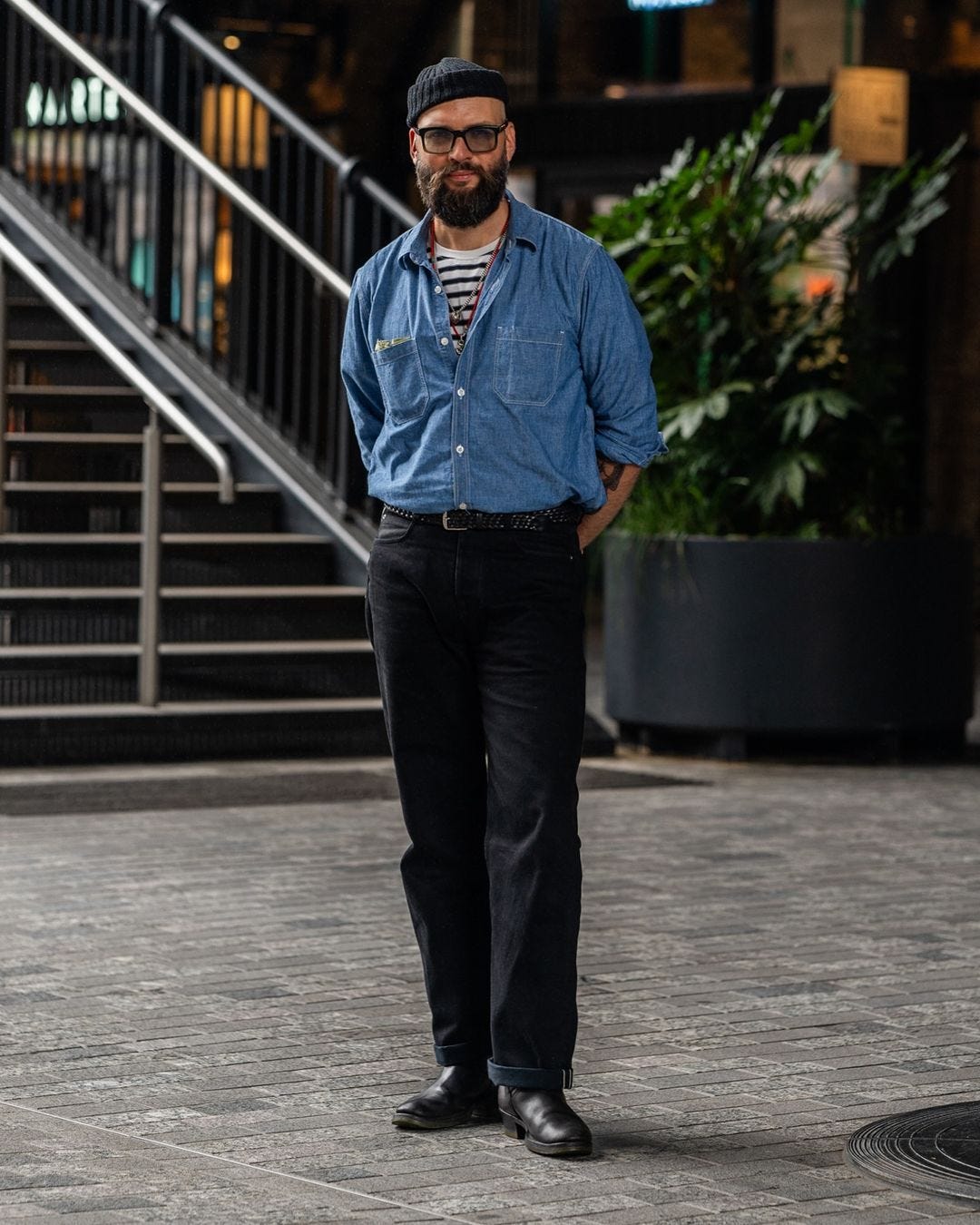 man standing in a lobby wearing a chambray shirt buttoned over a blue and white striped shirt with black pants