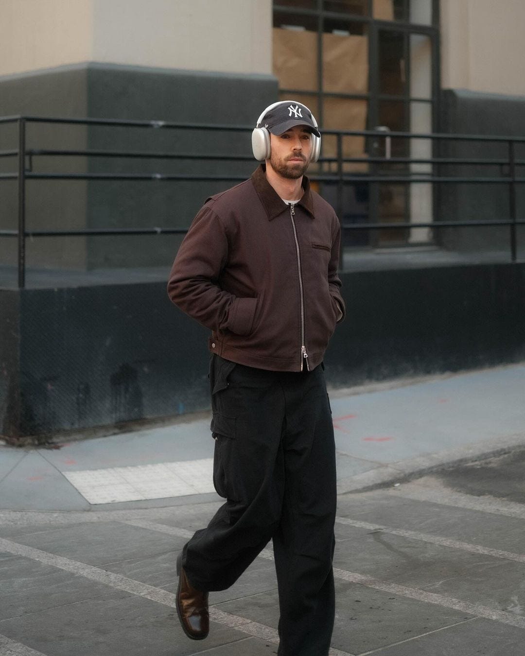 young man walking across the street wearing silver headphones, a brown workwear jacket and black pants. his hands are in his pockets