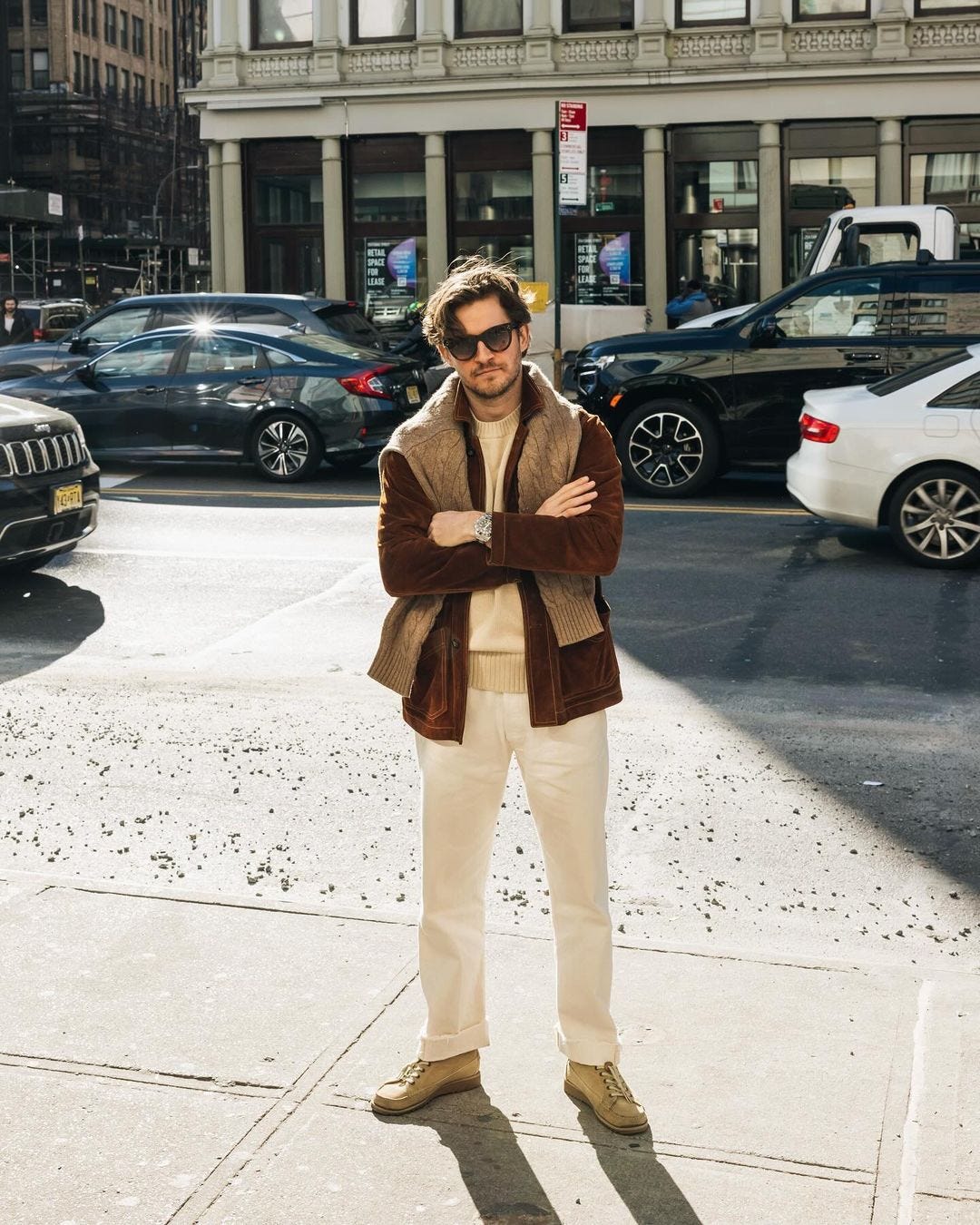 Man wearing sunglasses standing on sidewalk, wearing brown coat, tan sweater on shoulders, light pants and suede shoes