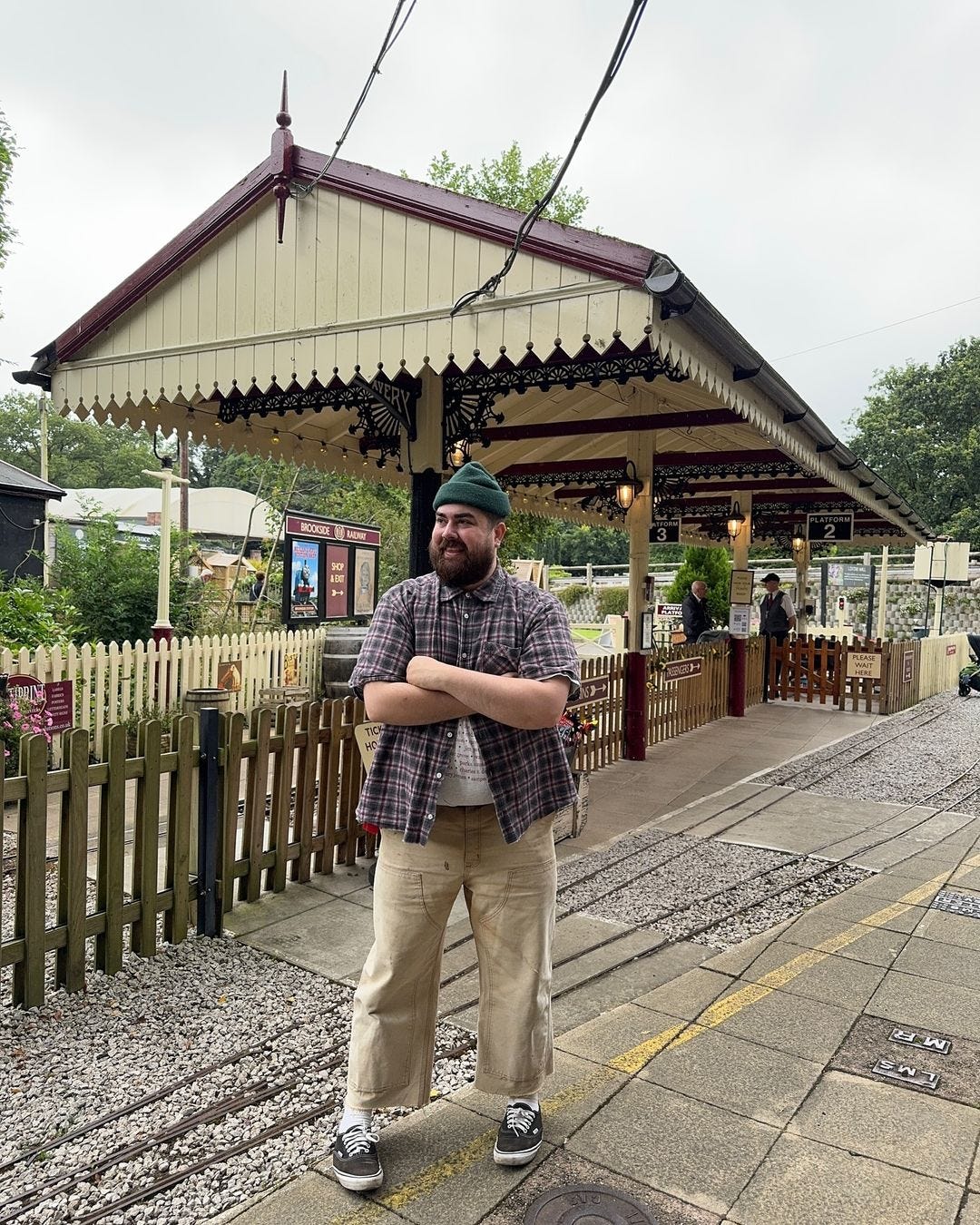 Man standing at train station wearing short-sleeved plaid shirt and ankle-baring khaki pants